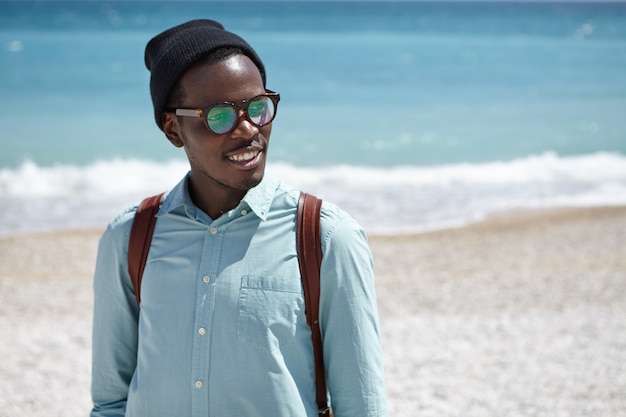 Cheerful young Afro American male student wearing eyeglasses and hat carrying knapsack on his shoulders spending leisure time after college by the sea, having nice walk along desert pebble beach