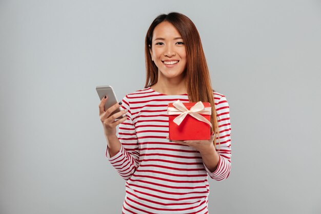 陽気な若いアジア女性がギフトを保持している電話でチャット。