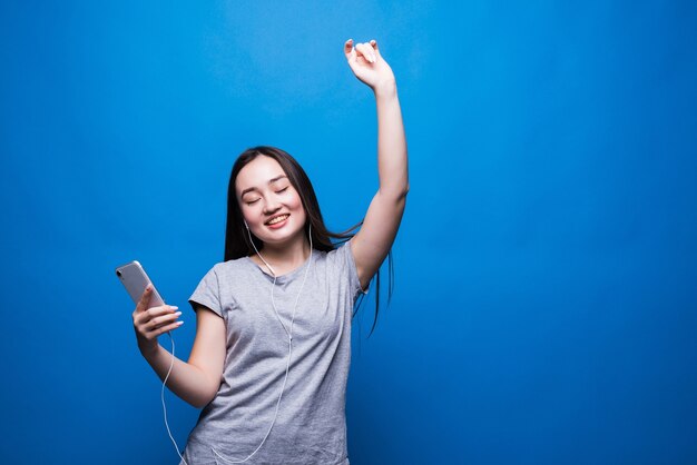 Веселая молодая азиатская женщина в наушниках, слушающая музыку и танцы, изолированные на синей стене