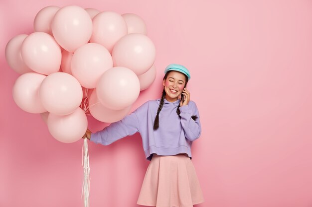Cheerful young Asian woman holds air balloons, calls friend via smartphone, gets pleasure to receive congratulation from close people, dressed in fashionable clothing.