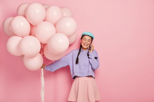 Жизнерадостная молодая азиатка держит воздушные шарики, звонит другу через смартфон, с удовольствием принимает поздравления от близких людей, одетых в модную одежду.