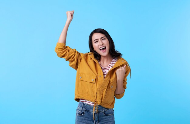 Cheerful young Asian woman raising his fists with smiling delighted face, yes gesture, celebrating success on blue background.