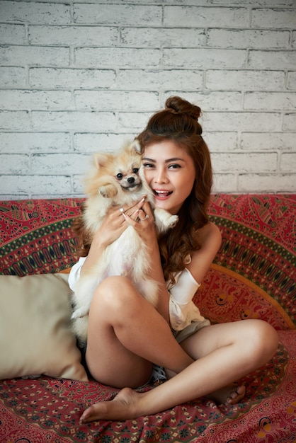 Cheerful young Asian woman sitting on couch at home and holding small dog