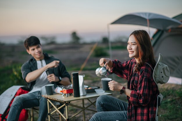 コーヒーセットと森のテントの前に座って、夏休みのキャンプ旅行中に新鮮なコーヒーグラインダーを作る陽気な若いバックパッカーカップル、選択的な焦点