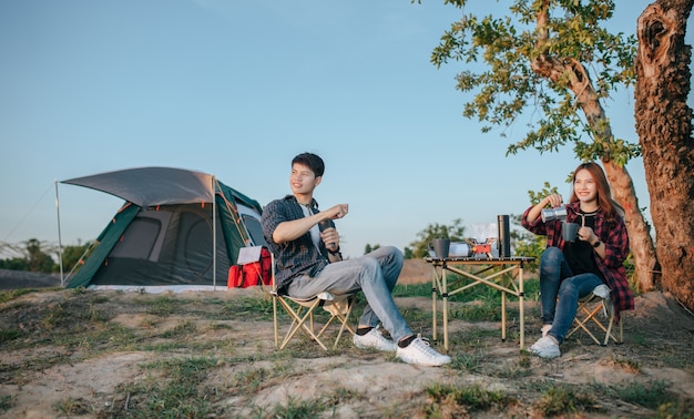 Веселая молодая пара пеших туристов сидит перед палаткой в лесу с кофейным сервизом и делает свежую кофемолку во время похода на летние каникулы