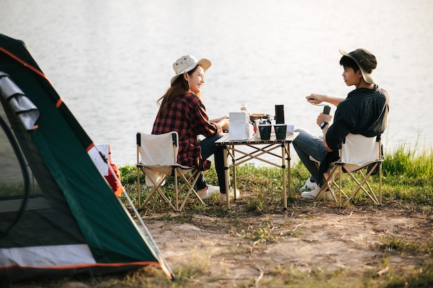 夏休みのキャンプ旅行中にコーヒーセットと淹れたてのコーヒーグラインダーを作る森のテントの前に座っている陽気な若いバックパッカーカップル