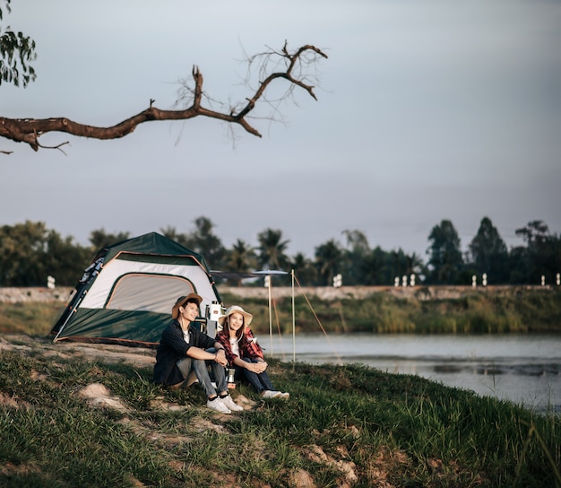 Веселая молодая пара пеших туристов сидит перед палаткой возле озера с кофейным сервизом и делает свежую кофемолку во время похода на летние каникулы