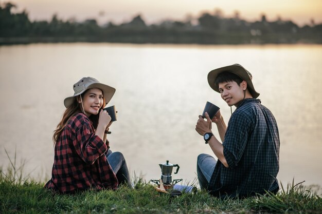 早朝に湖の近くの芝生に座って、夏休みのキャンプ旅行中に淹れたてのコーヒーグラインダーを作る陽気な若いバックパッカーカップル