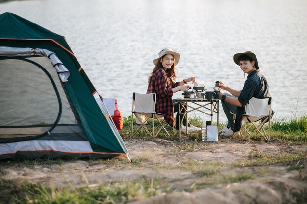 cheerful young backpacker couple wearing trekking hat sitting near lake with coffee and breakfast and making fresh coffee grinder while camping trip on summer vacation