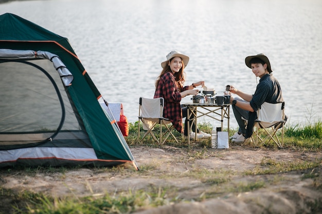 Веселая молодая пара пеших туристов в треккинговой шляпе сидит возле озера с кофе и завтраком и делает свежую кофемолку во время похода на летние каникулы