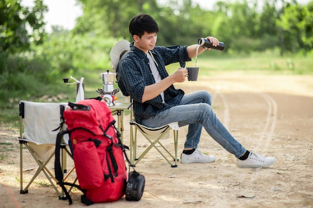 夏休みのキャンプ旅行中にコーヒーセットと淹れたてのコーヒーグラインダーを作る森のテントの前に座っている陽気な若いバックパッカーの男