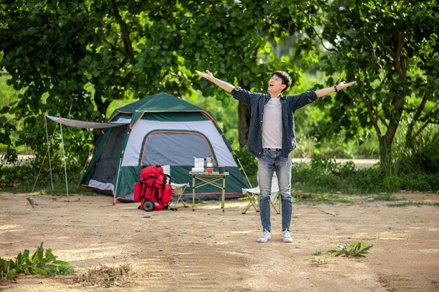 Cheerful Young backpacker man standing and open arms at front of the tent in forest with coffee set and making fresh coffee grinder while camping trip on summer vacation