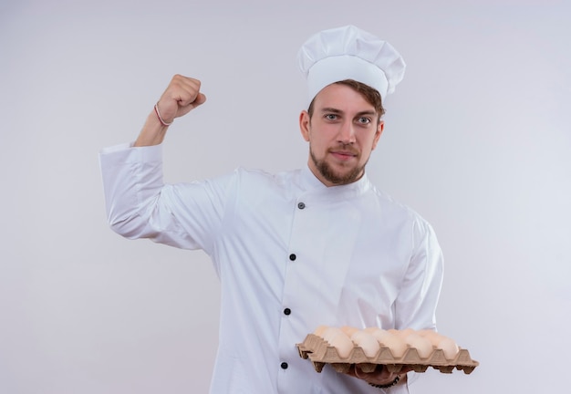 白い壁を見ながら、白い炊飯器の制服とくいしばられた握りこぶしで卵のカートンを保持している帽子を身に着けている陽気な若いひげを生やしたシェフの男