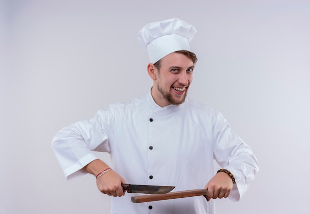 A cheerful young bearded chef man wearing white cooker uniform and hat holding knife over a wooden kitchen board while looking on a white wall