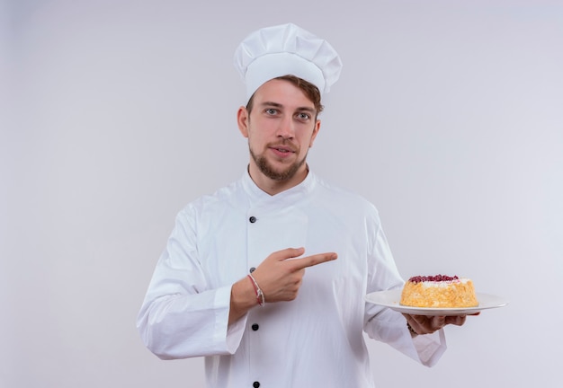 A cheerful young bearded chef man wearing white cooker uniform and hat pointing at a plate with cake with index finger while looking on a white wall