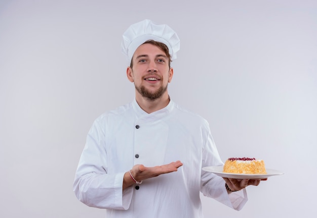 白い壁を見ながらケーキとプレートを示す白い炊飯器の制服と帽子を身に着けている陽気な若いひげを生やしたシェフの男