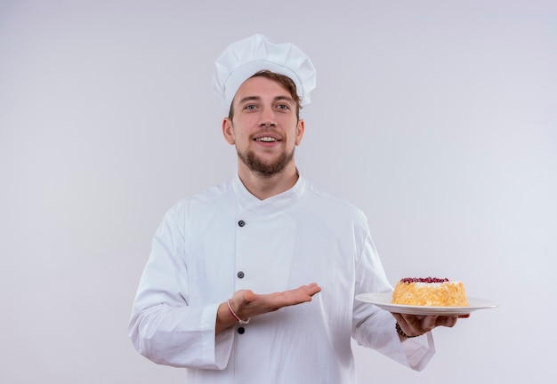 Веселый молодой бородатый повар в белой униформе и шляпе показывает тарелку с тортом, глядя на белую стену