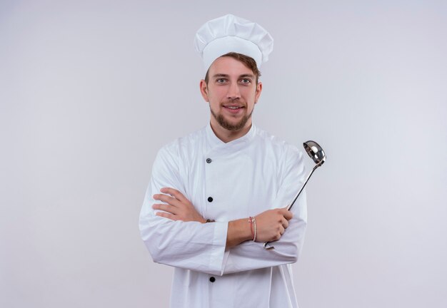 Un allegro giovane chef barbuto uomo in uniforme bianca tenendo il mestolo mentre guarda su un muro bianco