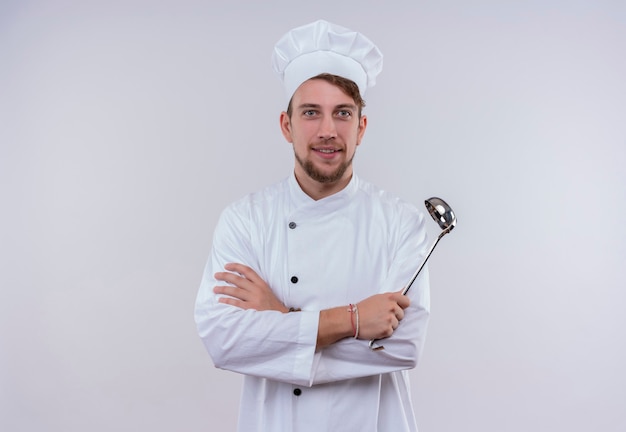 A cheerful young bearded chef man in white uniform holding ladle while looking on a white wall