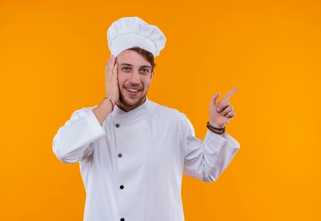 A cheerful young bearded chef man in white uniform pointing up with index finger while holding hand on face and looking on an orange wall