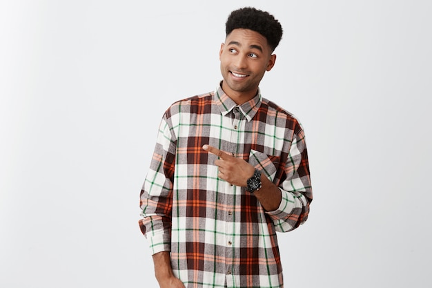 Cheerful young beautiful black man with afro hairstyle in stylish casual shirt smiling with teeth, pointing on free space, looking aside with excited and delight expression
