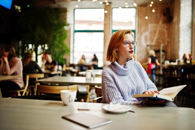 Веселая молодая красивая рыжеволосая женщина в очках сидит на своем рабочем месте в кафе и пьет кофе