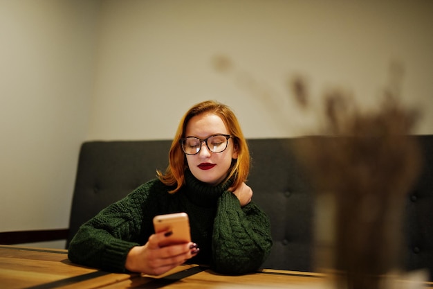 Веселая молодая красивая рыжеволосая женщина в очках в зеленом теплом шерстяном свитере сидит в кафе и пользуется мобильным телефоном