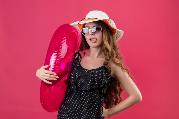 Веселая молодая красивая девушка-путешественница в платье в горошек в летней шляпе в солнцезащитных очках держит надувное кольцо, счастливая и позитивная, высунув язык, стоящий над розовым пространством