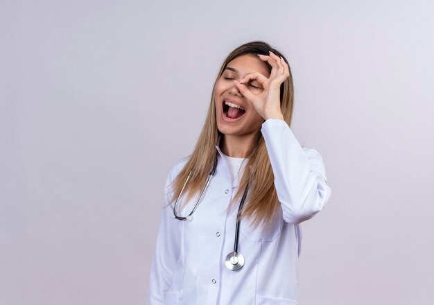 Cheerful young beautiful woman doctor wearing white coat with stethoscope doing ok sign with fingers looking through this sign