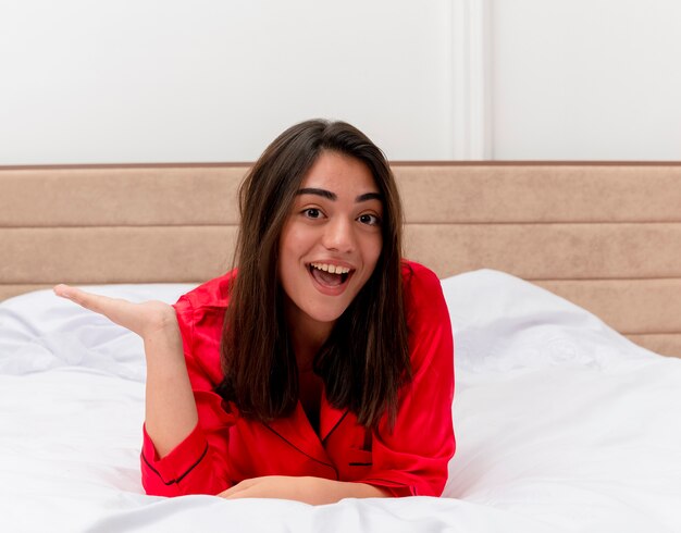 Веселая молодая красивая женщина в красной пижаме, лежа на кровати, счастлива и позитивно улыбается в домашнем интерьере на светлом фоне