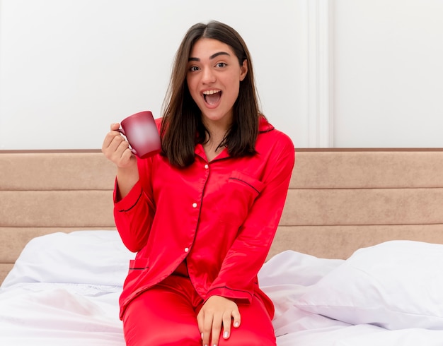 Cheerful young beautiful woman in red pajamas sitting on bed with cup of coffee looking at camera happy and excited in bedroom interior on light background