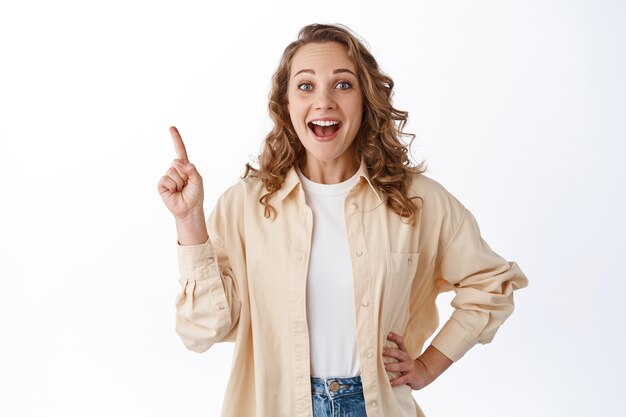Cheerful young blond girl with curly hair, stylish clothes, pointing finger at upper left corner, showing promo offer, standing over white wall