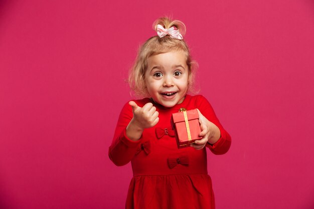 Cheerful young blonde girl in red dress holding gift box