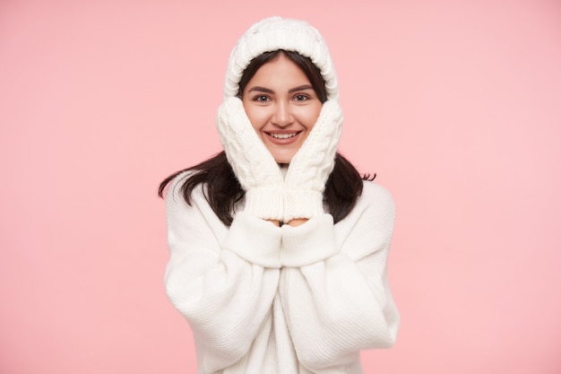 Cheerful young brown-eyed brunette woman dressed in white wollen cosy clothes holding her face with raised hands and smiling happily at front, isolated over pink wall