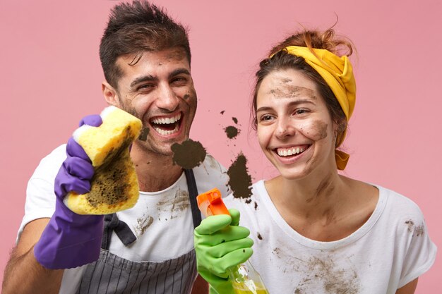 Веселая молодая кавказская пара с грязными лицами вместе убирают дом. Улыбающаяся симпатичная женщина и ее муж, оба в защитных перчатках, моют окно с помощью спрея и губки