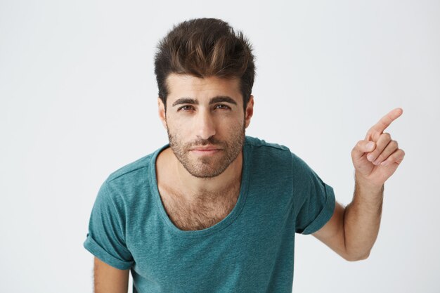 Cheerful young Caucasian male wearing blue t-shirt with stylish haircut pointing his index finger sideways at blank white copy space wall, indicating something extraordinary