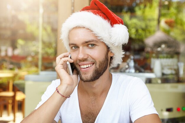 전화 대화를 나누는 산타 클로스 모자에 쾌활 한 젊은 백인