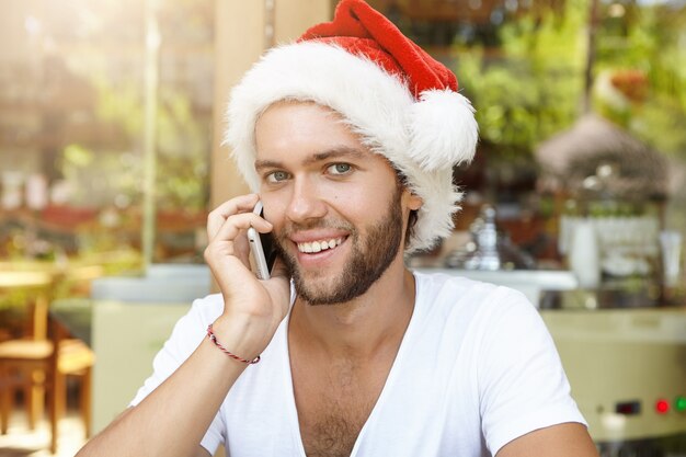 Cheerful young Caucasian in Santa Claus hat having phone conversation