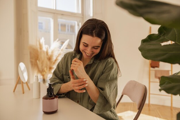 Cheerful young caucasian woman applies cream on her hands while sitting at table in light room Brunette girl with smooth skin wears shirt Wellness and self care concept