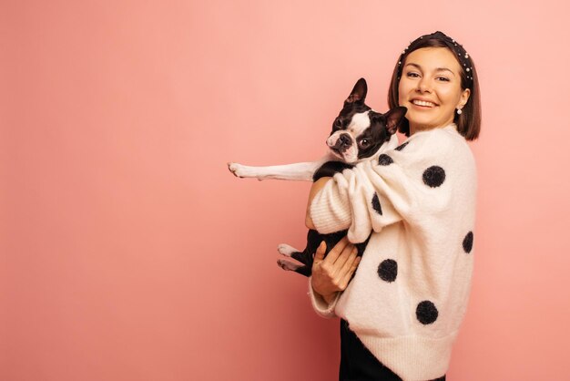 Cheerful young caucasian woman holding black and white bulldog while standing on pink background with space for text Love between dog and owner concept