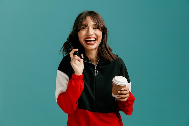 Веселая молодая кавказская женщина, держащая мобильный телефон возле уха и чашку кофе на вынос, смотрит в камеру и смеется на синем фоне