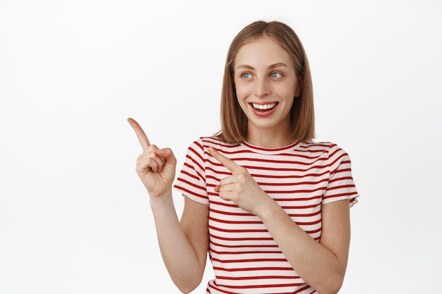 Cheerful young caucasian woman with blond hair, laughing and smiling happy, pointing fingers left, looking at sale promo text, showing advertisement, white background.