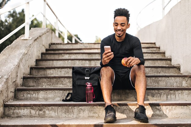 Веселый молодой крутой темнокожий спортсмен в черных шортах и футболке сидит на лестнице, улыбается и держит яблоко и телефон