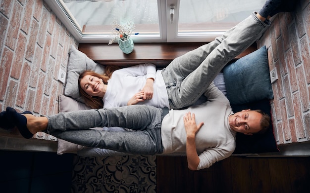 Free photo cheerful young couple lying on window seat at home