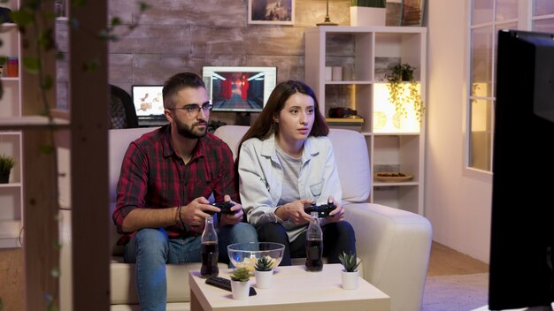 쾌활한 젊은 부부는 소파에 앉아서 텔레비전에서 비디오 게임을 하고 있습니다. 행복한 관계