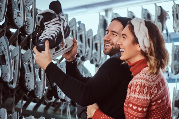 Cheerful young couple wearing warm clothes standing near rack with many pairs of skates, laughing while choosing ice skates