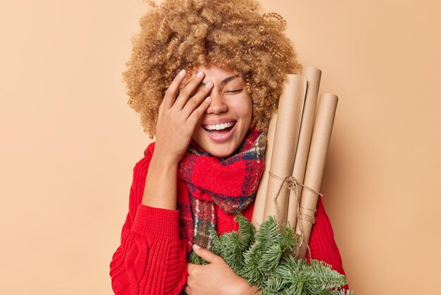 陽気な若い縮れ毛の女性は、飾りの枝と紙のローラーを持ってクリスマスや新年を待っていますベージュの背景の上に隔離された暖かい冬の服を着た顔の手のひらを作ります