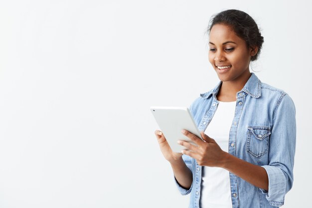 Cheerful young dark-skinned female student with cute smile standing on white wall, using tablet, checking newsfeed on her social network accounts. Pretty afro-american girl surfing internet on t