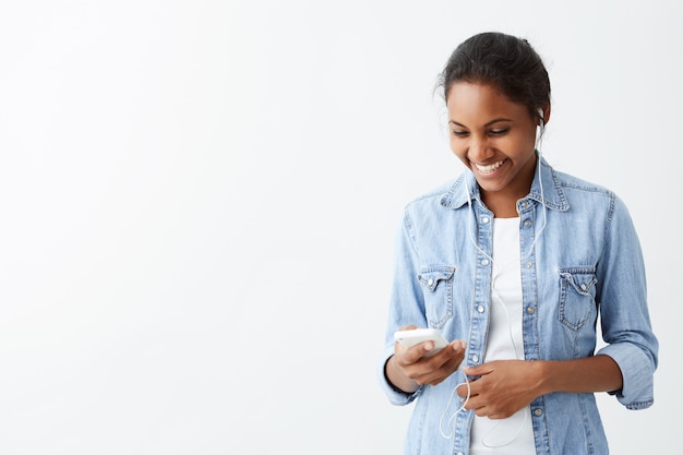 Веселая молодая темнокожая молодая женщина в синей рубашке, чувствуя себя счастливой и взволнованной, читая сообщение на смартфоне, получая некоторые позитивные новости