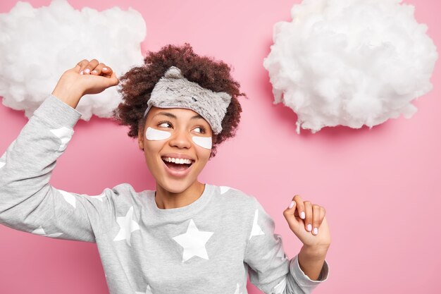 Cheerful young ethnic teenage girl smiles broadly dances and moves actively raises arms has good mood in morning wears comfortable home wear isolated over pink wall clouds above